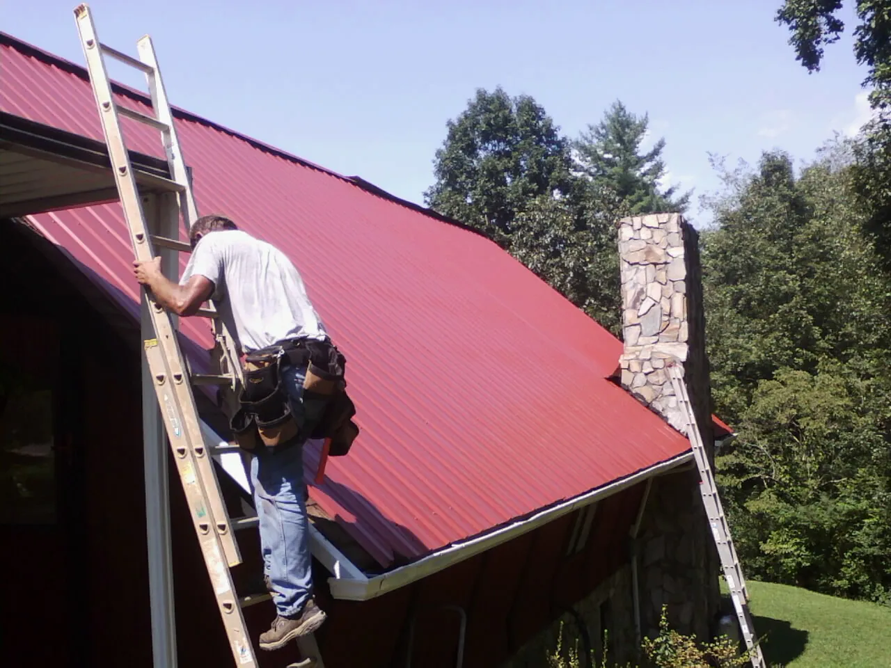 Roofer on a ladder beside a red metal roof and stone chimney — Soffit & Fascia Repair by Roofing Brothers in Ronkonkoma